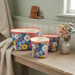 Peut inclure: Trois paniers de rangement en tissu de tailles variées présentant un fond bleu vif avec un motif floral coloré composé de fleurs jaunes, rouges et blanches. Chaque panier possède un bord corail uni et une doublure intérieure de couleur crème.