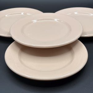 May include: A set of three round, light beige ceramic plates with a smooth, glossy finish. The plates feature a wide, flat rim and a slightly recessed center. They are arranged in a triangular formation, with one plate positioned in the foreground.