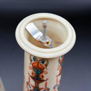 May include: Close-up of a vintage ceramic pepper grinder. The top has a cream-coloured rim and a metal grinding mechanism. The body of the grinder is decorated with a colourful, ornate design featuring faces and floral elements.