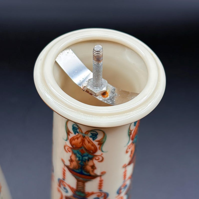 May include: Close-up of a vintage ceramic pepper grinder. The top has a cream-coloured rim and a metal grinding mechanism. The body of the grinder is decorated with a colourful, ornate design featuring faces and floral elements.