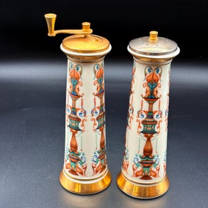 May include: A pair of decorative ceramic salt and pepper grinders. The grinders have a cream-coloured base with ornate red, teal, and gold floral designs. The tops and bases are gold-coloured, with one grinder featuring a silver-coloured top.