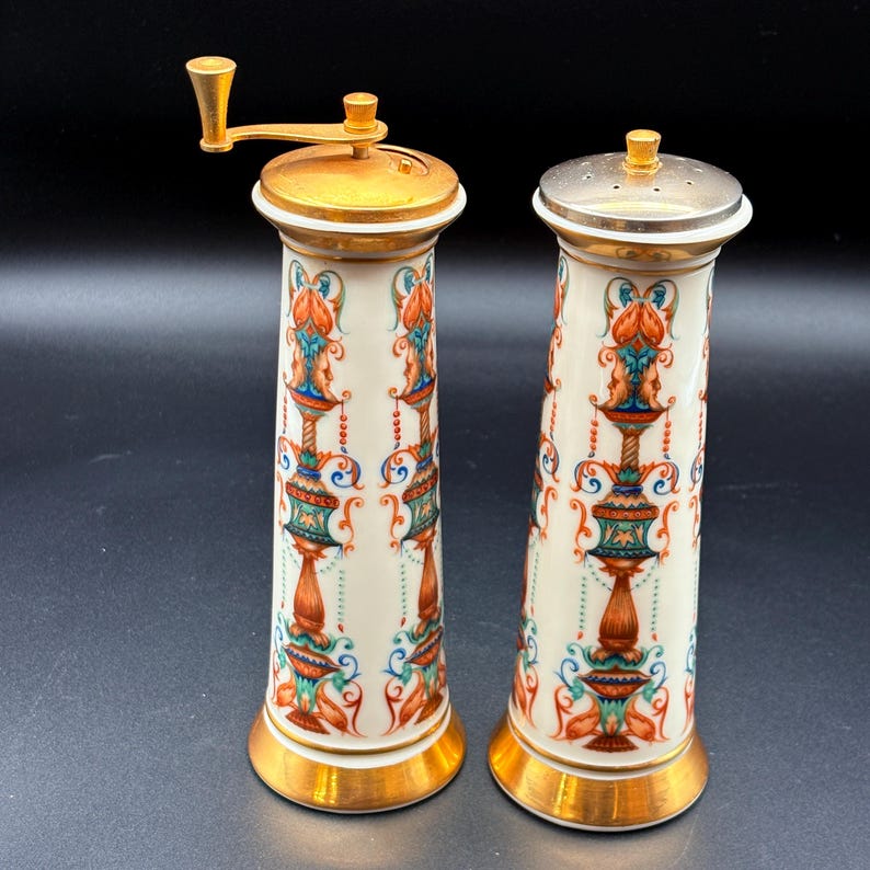 May include: A pair of decorative ceramic salt and pepper grinders. The grinders have a cream-coloured base with ornate red, teal, and gold floral designs. The tops and bases are gold-coloured, with one grinder featuring a silver-coloured top.