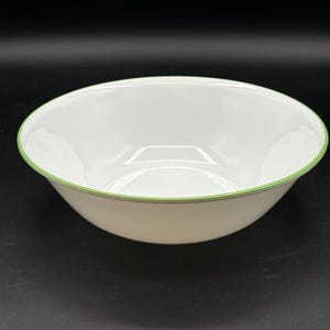 May include: White ceramic bowl with a light green trim around the rim. The bowl has a simple, classic design and is set against a dark background. The bowl is likely used for serving food.