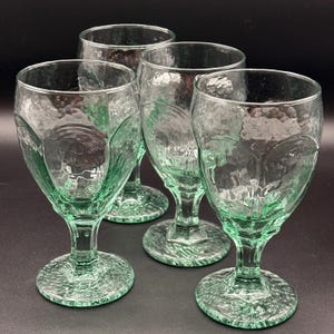 May include: Four green-tinted water goblets with textured surfaces. The glasses have a unique design with a textured pattern on the bowl and base. The stemware is arranged on a dark surface, showcasing the glassware's details.