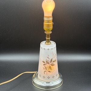 May include: A vintage table lamp with a frosted glass shade featuring gold floral designs. The lamp has a brass neck and a round, silver-coloured base. The bulb is lit, emitting a warm glow. The lamp's cord is a light yellow colour.