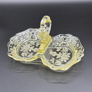 Puede incluir: Un plato de servir de vidrio amarillo con asa y dos compartimentos. El plato presenta un diseño floral y un borde festoneado. El asa es arqueada y une las dos secciones del plato.