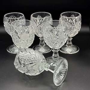 May include: A set of six clear crystal wine glasses. Each glass features a diamond-cut pattern on the bowl and a stemmed base. One glass is lying on its side. The glasses are arranged on a black surface.