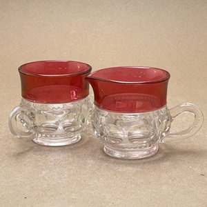 Puede incluir: Un juego de dos jarras de crema y azúcar de vidrio transparente con un borde rojo. Las jarras tienen un patrón decorativo.