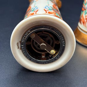 May include: Close-up of a white ceramic salt or pepper grinder with a metal grinding mechanism. The base reads "Mayfair Products Inc. Trenton, N.J." and "Lenox". The top has a colorful floral design with gold accents.