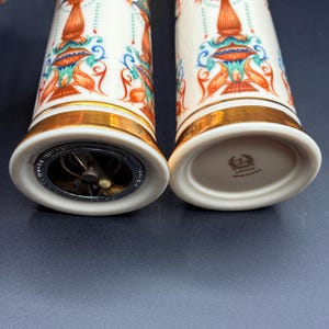 May include: A pair of white ceramic salt and pepper shakers with gold bands. They feature a colourful design with orange, blue, and green details. One shaker has a metal grinder at the base, the other has the Lenox logo. The shakers are for seasoning food.