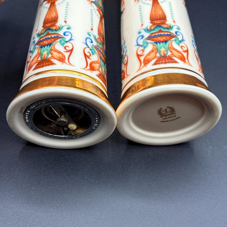 May include: A pair of white ceramic salt and pepper shakers with gold bands. They feature a colourful design with orange, blue, and green details. One shaker has a metal grinder at the base, the other has the Lenox logo. The shakers are for seasoning food.
