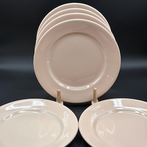May include: A set of five smooth, pale pink ceramic plates. Three plates are positioned in the foreground, while two are stacked vertically behind them, supported by a small wooden stand. The plates feature a simple, circular design.