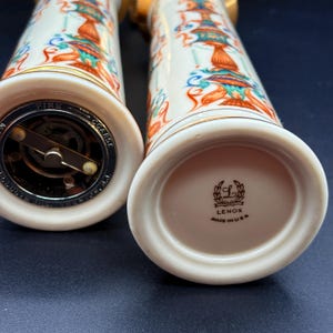 May include: Two white Lenox salt and pepper shakers with a colourful design of orange, teal, and blue figures. The shakers have gold trim and metal grinding mechanisms. The base of one shaker reads "Lenox Made in USA."