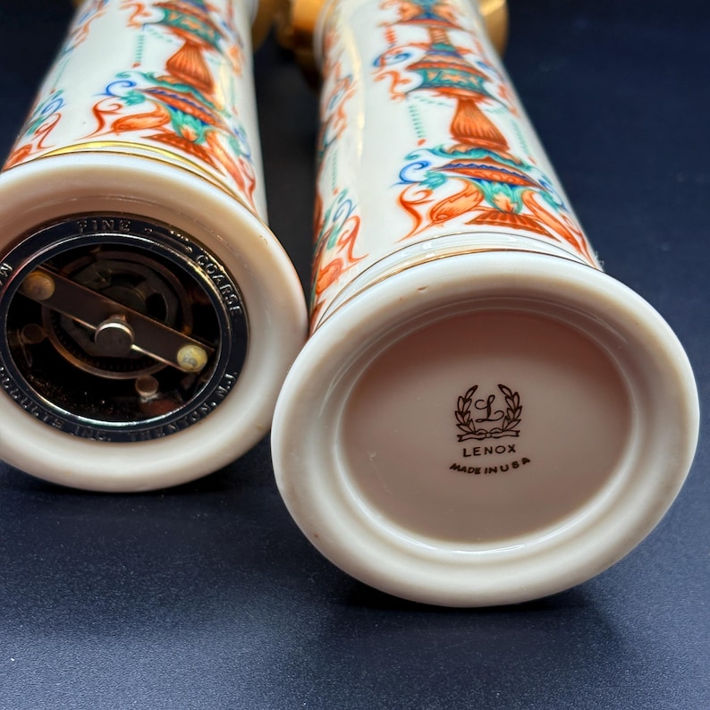 May include: Two white Lenox salt and pepper shakers with a colourful design of orange, teal, and blue figures. The shakers have gold trim and metal grinding mechanisms. The base of one shaker reads "Lenox Made in USA."