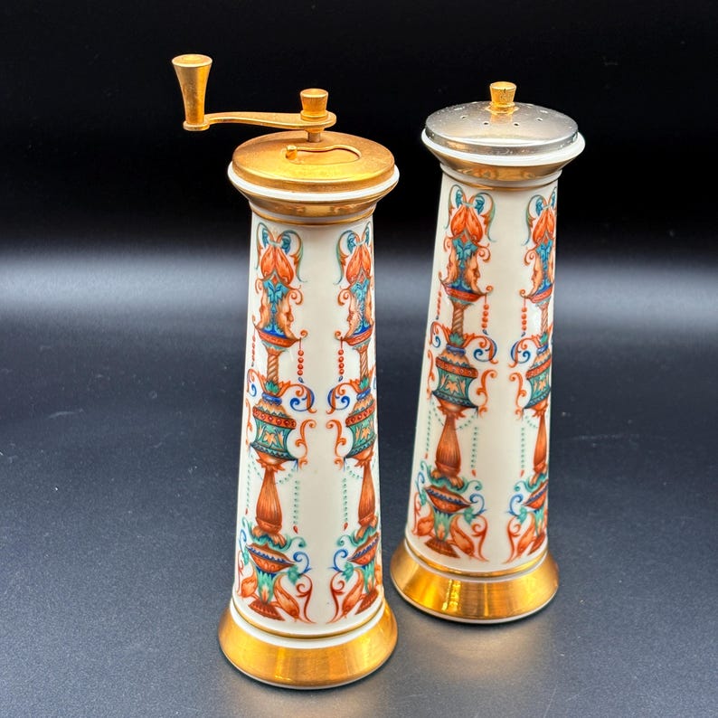 May include: A pair of antique ceramic salt and pepper shakers. The shakers are white with a repeating pattern of orange, blue, and green floral designs. They have gold-coloured accents on the top and bottom. One shaker has a metal grinder, and the other has a metal top.