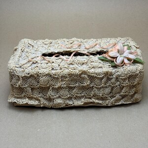 May include: A cream-coloured, rectangular tissue box cover, made with a detailed crochet lace design. It has a rectangular opening on top and is decorated with a pink flower and ribbon. The style is vintage and decorative, suitable for a bedroom or bathroom.