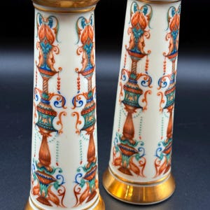 May include: Two white porcelain vases with ornate, colourful designs. The vases feature gold rims and bases, with detailed patterns in orange, teal, blue, and gold. The design includes stylized faces and floral motifs.