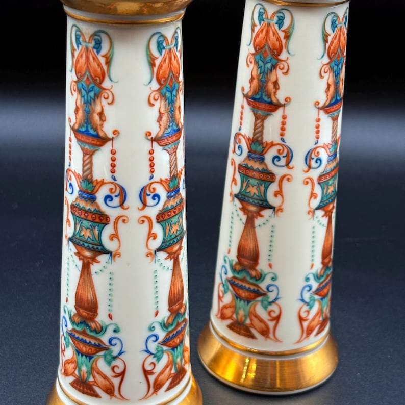May include: Two white porcelain vases with ornate, colourful designs. The vases feature gold rims and bases, with detailed patterns in orange, teal, blue, and gold. The design includes stylized faces and floral motifs.