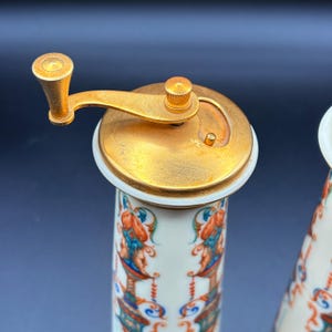 May include: A close-up of a vintage pepper grinder with a gold-coloured metal top and handle. The ceramic body is white with a colourful floral and figure design. A classic kitchen accessory.