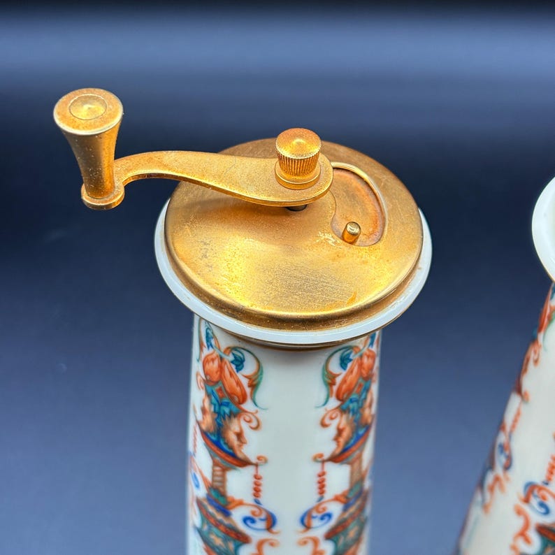 May include: A close-up of a vintage pepper grinder with a gold-coloured metal top and handle. The ceramic body is white with a colourful floral and figure design. A classic kitchen accessory.