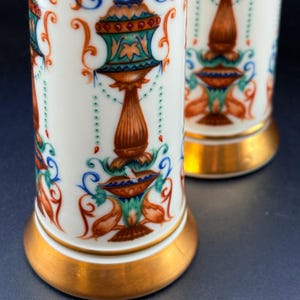 May include: Two white porcelain vases with ornate, colourful designs. The vases have a gold base and are decorated with orange, blue, and green floral patterns. The design includes a central, brown, pedestal-like shape with a detailed top.