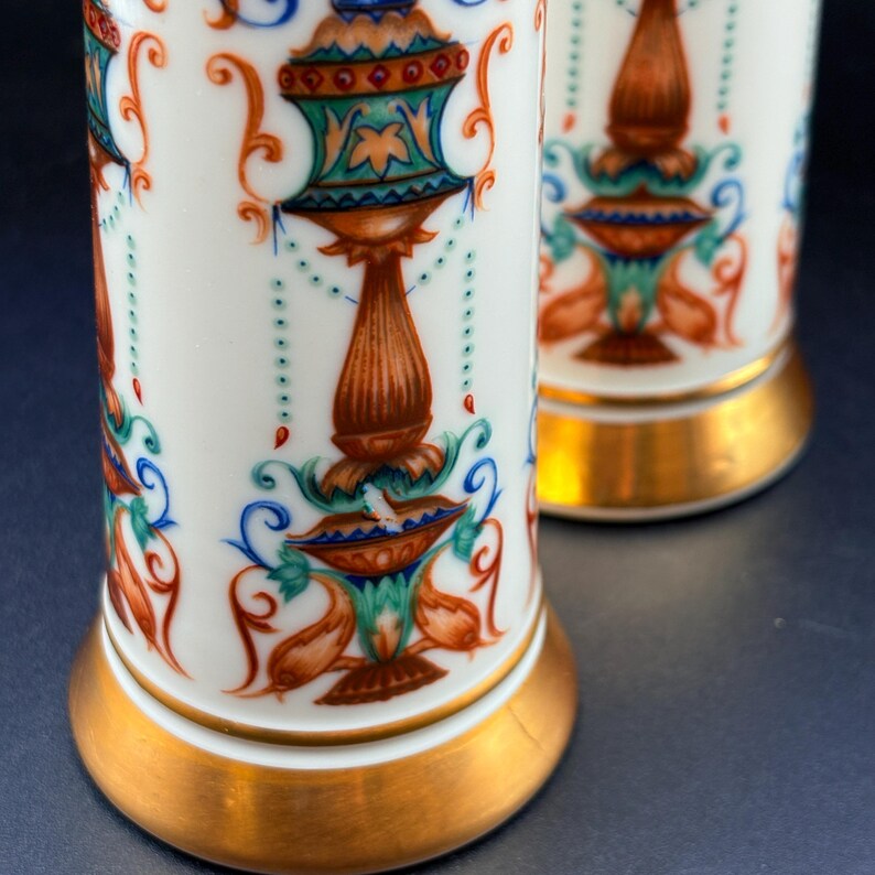 May include: Two white porcelain vases with ornate, colourful designs. The vases have a gold base and are decorated with orange, blue, and green floral patterns. The design includes a central, brown, pedestal-like shape with a detailed top.