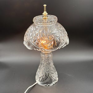 May include: A clear, cut-glass table lamp with a decorative pattern. The lamp features a mushroom-shaped shade, a pedestal base, and a gold-colored finial. The lamp is lit, casting a warm glow.