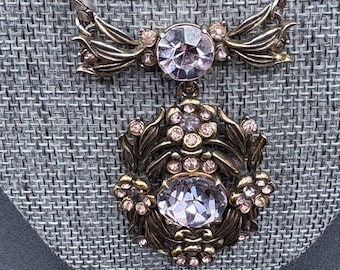 May include: Vintage necklace with a silver-tone chain and a pendant featuring a large, clear, faceted stone. The pendant is surrounded by an ornate, bronze-coloured metal design with smaller pink stones. The necklace has a decorative bow-shaped element.