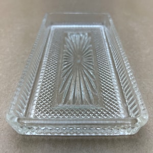 Puede incluir: Una bandeja rectangular de vidrio transparente con un patrón decorativo en forma de estrella en el centro. La bandeja tiene un borde elevado y texturizado.