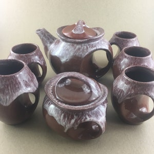 May include: A brown and white ceramic teapot and four matching mugs with unique handles. The teapot has a lid and a spout. The mugs have a brown glaze with white speckles. The set is a vintage style.