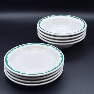 May include: A set of white ceramic bowls and plates with a green decorative border. The bowls are stacked in two groups, with a stack of bowls and a stack of plates. The plates are flat with a slightly raised edge.