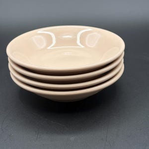 May include: A stack of four round, light beige ceramic bowls with a smooth, glossy finish. Each bowl features a shallow basin and a slightly curved rim, creating a uniform appearance. The stack is arranged vertically, showcasing the consistent color.