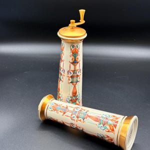 May include: Two decorative ceramic pepper grinders. The grinders have a cream-coloured base with ornate, colourful patterns in orange, teal, and blue. The tops and bottoms are trimmed with gold. One grinder is upright, and the other is lying down.