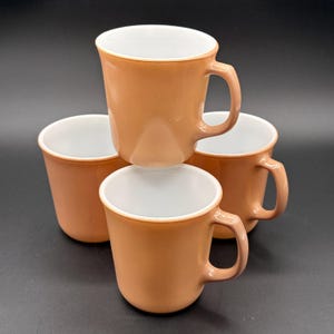 Puede incluir: Cuatro tazas de cerámica con un exterior marrón anaranjado cálido y un interior blanco brillante. Cada taza tiene un asa curva y un borde ligeramente ensanchado. Las tazas están apiladas sobre un fondo oscuro.