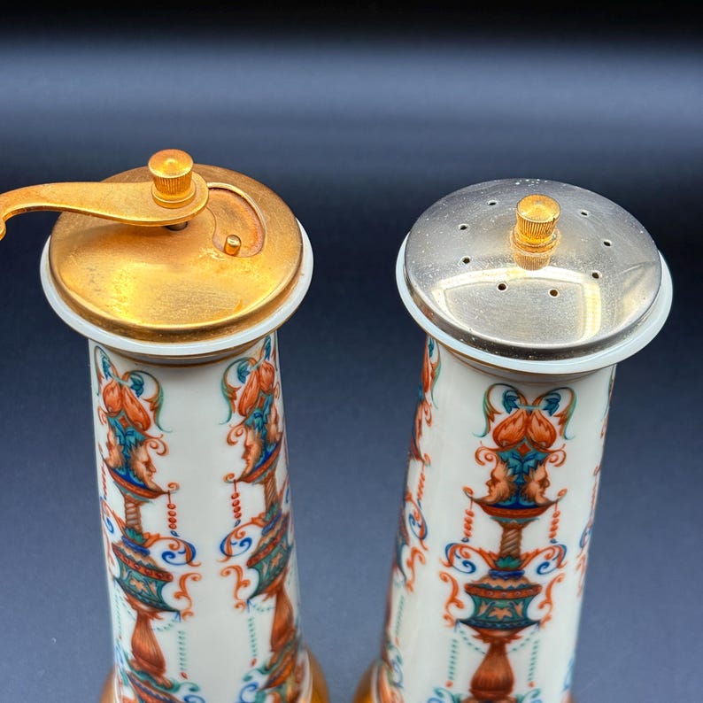 May include: A pair of decorative salt and pepper shakers. The shakers have a white base with a colourful design of faces and floral patterns. The salt shaker has a silver top with small holes, and the pepper shaker has a gold-coloured grinder.
