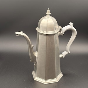 May include: A vintage silver-colored coffee pot with a faceted body and a curved spout. The pot has a lid with a small finial and a decorative handle. The design is reminiscent of antique kitchenware.