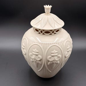 May include: Cream-colored ceramic jar with a lid. The jar has a rounded shape with embossed floral designs and vertical ribbed detailing. The lid features a floral finial and a decorative pierced design. The jar is on a dark background.