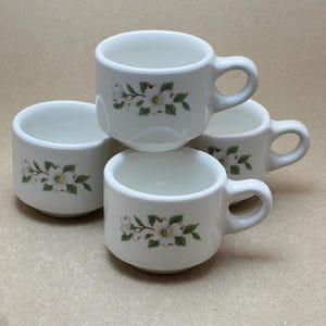 May include: Four white ceramic coffee mugs with a floral design of white flowers and green leaves. The mugs are stacked on top of each other.