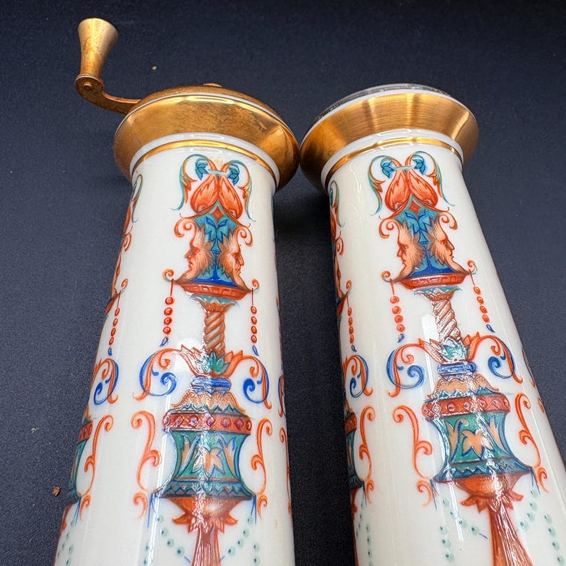 May include: Two white ceramic salt and pepper grinders with gold-coloured tops and handles. The grinders feature ornate, colourful designs with faces and floral patterns. The design includes orange, blue, and green accents.