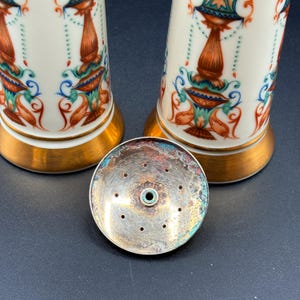 May include: Two white ceramic salt and pepper shakers with detailed, colourful designs. The shakers have gold-coloured bases and are decorated with orange, blue, and green floral patterns. A silver-coloured lid with holes is in front.