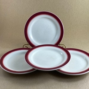 May include: Four white ceramic plates with a burgundy rim. The plates are arranged on a gold-colored metal stand. The plates are oval and round.