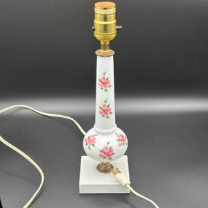 May include: A vintage white table lamp with pink floral designs. The lamp has a brass base and a square marble base. The lamp is approximately 12 inches tall.