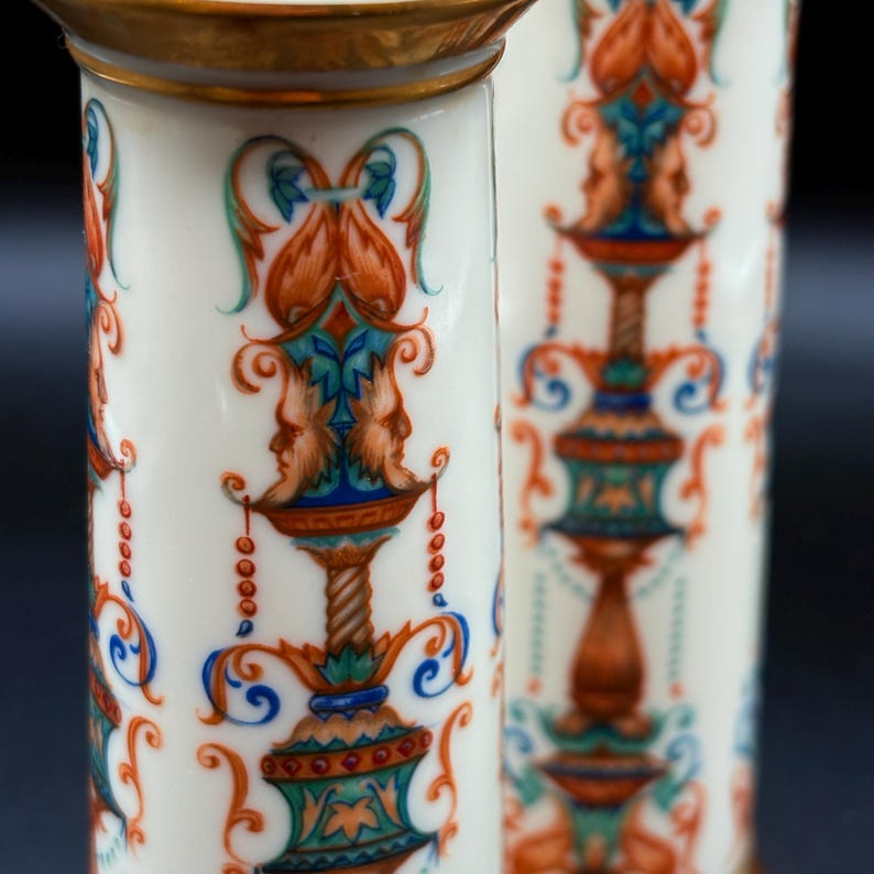 May include: Two white ceramic columns with intricate, colourful designs. The columns display a repeating pattern of orange, blue, and green floral and face motifs. Gold trim adorns the top of each column, creating a luxurious aesthetic.