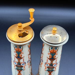 May include: A pair of vintage ceramic salt and pepper shakers. The salt shaker has a silver top with small holes, while the pepper grinder has a gold-coloured crank. Both shakers are white with a colourful floral and face design.