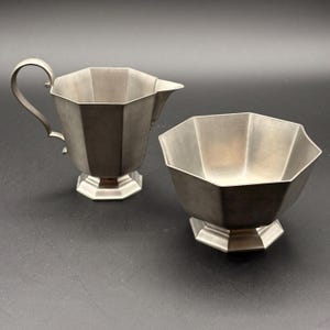 Può includere: Un set vintage di crema e zuccheriera in peltro. I pezzi hanno un design geometrico sfaccettato e poggiano su basi ottagonali. La crema ha un manico curvo e un beccuccio. La zuccheriera è aperta.