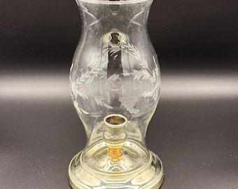 May include: A clear glass candle holder with a silver base and a gold-colored candle holder. The glass has an etched floral design. The candle holder is designed to hold a single candle.
