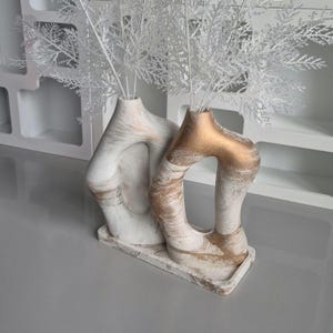 May include: Two abstract white vases with gold accents, each holding white decorative branches. The vases have a unique, open design and sit on a matching rectangular base. The background features a white shelf.