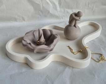 May include: A cream-colored, cloud-shaped tray with a gray ring holder, a small sculpture of a person, and a gold bracelet. The ring holder is shaped like a flower. A gold ring is in the ring holder. The sculpture is of a person sitting on a ball.