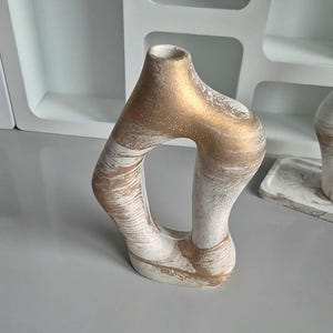 May include: A decorative ceramic vase with a unique, open design. The vase features a white base with gold accents and a textured finish. The top and interior of the vase are gold. The vase is a modern home decor item.
