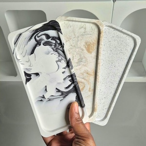 Peut inclure: Trois plateaux rectangulaires avec des bords surélevés sont présentés. Le premier est blanc avec un motif marbré noir et blanc, le second beige et or, et le troisième gris moucheté. Une main tient les plateaux, mettant en valeur leurs motifs décoratifs.